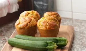 Vier goldbraune Zucchini-Muffins auf einem Holzbrett, daneben zwei frische Zucchini.