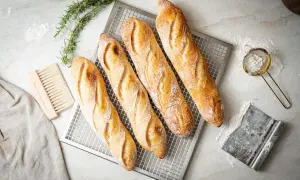 Vier frisch gebackene Baguettes auf einem Gitter, bereit für das Baguette Rezept. Daneben liegen ein Bürstchen, ein Tuch und ein Sieb mit Mehl.