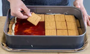 Eine Hand legt einen Butterkeks auf eine Schicht Marmelade und Creme in einer Backform. Der Butterkeks Kuchen wird geschichtet.