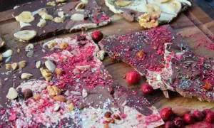 Bruchschokolade selber machen: Bunte Schokoladenstücke mit Nüssen, Beeren und Bananenchips auf Holzbrett.