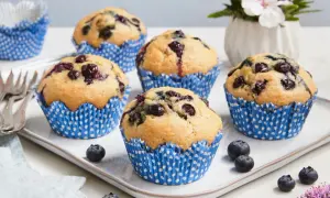 Fünf saftige, vegane Blaubeermuffins in blauen Papierförmchen auf einem Teller, garniert mit frischen Blaubeeren und einer Blume.