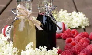 Zwei Flaschen mit selbstgemachtem Sirup, umgeben von Holunderblüten und Himbeeren. Perfekt zum Sirup einkochen.