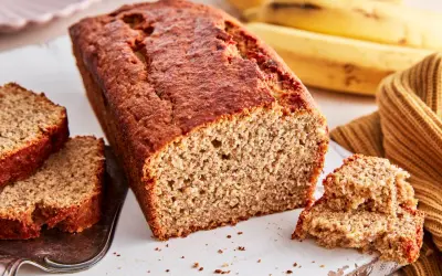 Ein saftiges, goldbraunes bananenbrot glutenfrei, mit zwei Scheiben auf einem Silbertablett und reifen Bananen im Hintergrund.