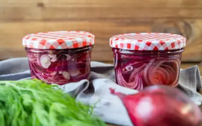 Zwei Gläser mit eingelegten roten Zwiebeln, bereit für Salate oder Sandwiches. Frischer Dill liegt daneben.