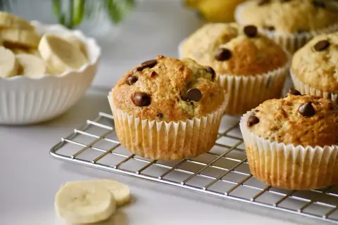 Frisch gebackene schoko bananen muffins mit Schokostückchen auf einem Kuchengitter. Daneben eine Schüssel mit Bananenscheiben.