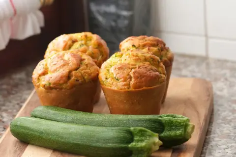 Vier goldbraune Zucchini-Muffins auf einem Holzbrett, daneben zwei frische Zucchini.