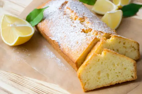 Saftiger veganer Zitronenkuchen, bestäubt mit Puderzucker, mit frischen Zitronenspalten und Blättern dekoriert.