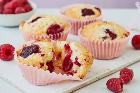 Saftige Himbeer Muffins in rosa Förmchen, einer aufgeschnitten, mit frischen Himbeeren garniert.