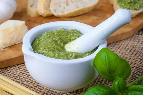 Frisches Pesto in einem Mörser, bereit zum Haltbarmachen mit Salz. Daneben liegen Brot, Parmesan, Knoblauch und Pasta.