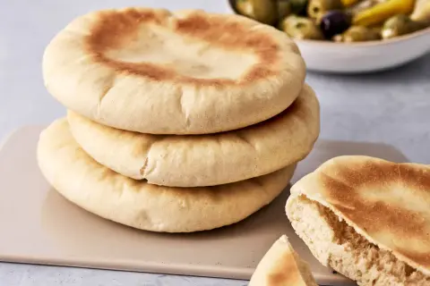Gestapeltes pita brot mit einer Schüssel Oliven im Hintergrund. Ein Stück ist aufgeschnitten und zeigt die weiche Krume.