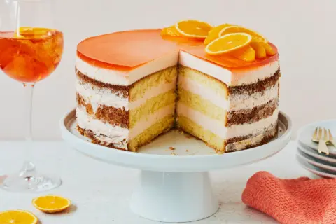 Eine Aperol Torte mit fruchtiger Geleehaube und Orangenscheiben, daneben ein Aperol Spritz.