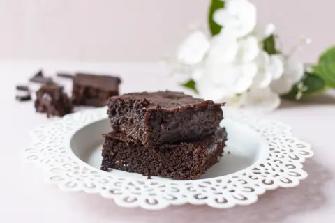 Zwei gestapelte, saftige low carb brownies auf einem weißen Teller, daneben zerbrochene Schokostücke und im Hintergrund weiße Blumen.