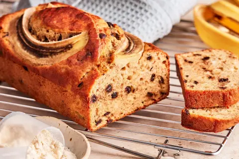Saftiges protein bananenbrot mit Schokostückchen und Bananenscheiben, daneben eine Schüssel mit Proteinpulver.