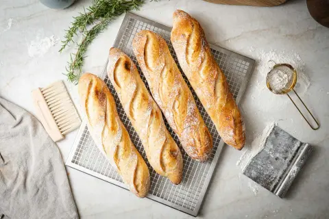 Vier frisch gebackene Baguettes auf einem Gitter, bereit für das Baguette Rezept. Daneben liegen ein Bürstchen, ein Tuch und ein Sieb mit Mehl.