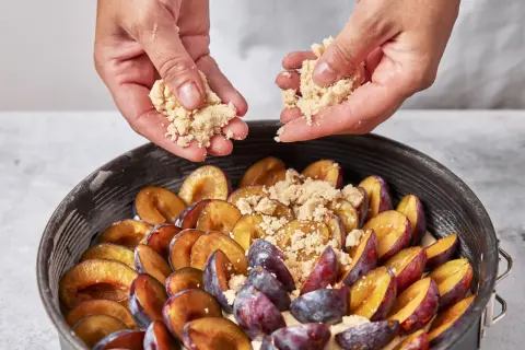Hände streuen Streusel über saftige Pflaumen in einer Springform. Der pflaumenkuchen nimmt Gestalt an.