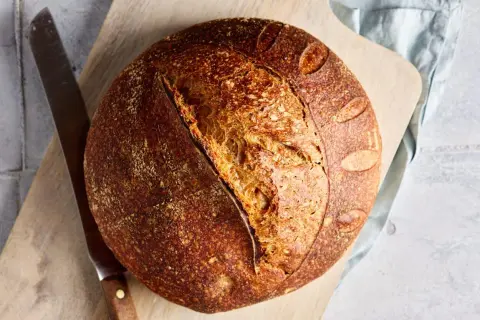 Ein rustikales Dinkel Sauerteigbrot mit knuspriger Kruste und luftiger Krume liegt auf einem Holzbrett neben einem Messer.