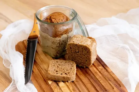 Brot im Glas Rezept: Ein Laib Brot im Glas, daneben ein Stück und ein kleines Quadrat auf einem Holzbrett. Ein Messer liegt daneben.