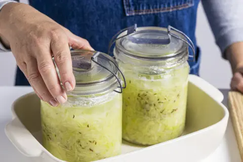 Hände setzen zwei Gläser mit Sauerkraut zum Fermentieren in ein Gefäß.