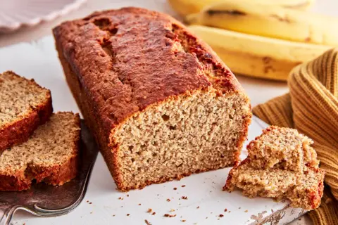 Ein saftiges, goldbraunes bananenbrot glutenfrei, mit zwei Scheiben auf einem Silbertablett und reifen Bananen im Hintergrund.