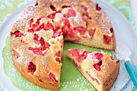 Ein saftiger Erdbeer Rhabarber Kuchen, bestäubt mit Puderzucker, liegt auf einem Teller. Ein Stück ist bereits herausgeschnitten.