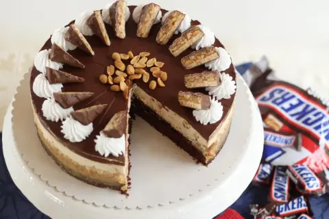 Eine köstliche Snickers Torte mit Schokoladenglasur, Erdnüssen und Sahnetuffs. Kleine Snickers-Stücke zieren den Rand.