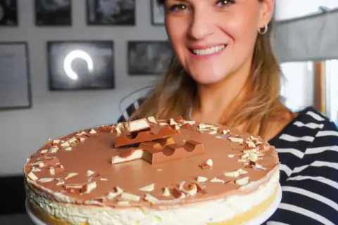 Lächelnde Frau präsentiert eine köstliche Kinderriegel Torte, dekoriert mit Schokostückchen und ganzen Riegeln.