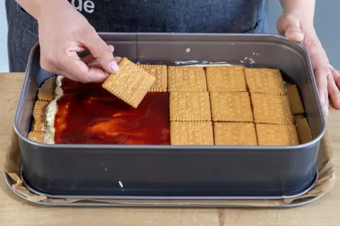 Eine Hand legt einen Butterkeks auf eine Schicht Marmelade und Creme in einer Backform. Der Butterkeks Kuchen wird geschichtet.