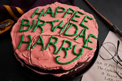 Ein Harry Potter Kuchen mit rosa Zuckerguss und grüner Schrift "Happy Birthday Harry". Daneben liegen eine Brille und ein Zauberstab.