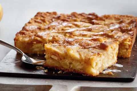 Saftige schwedische Apfeltorte, mit Äpfeln belegt und mit Puderzucker bestäubt, auf einem dunklen Brett mit einer Gabel.