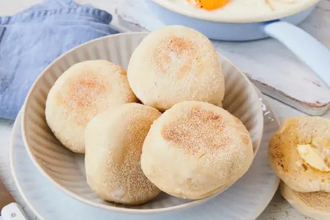 Ein Teller mit vier goldbraunen englischen Muffins, daneben ein halber, mit Butter bestrichener Muffin. Ein Spiegelei ist im Hintergrund zu sehen.