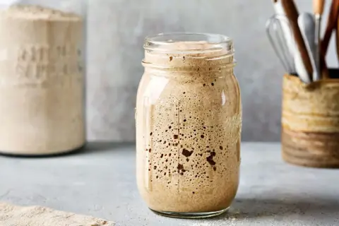 Ein Glas mit sprudelndem Sauerteig, bereit zum Backen. Im Hintergrund ein weiteres Glas mit Mehl und Küchenutensilien.