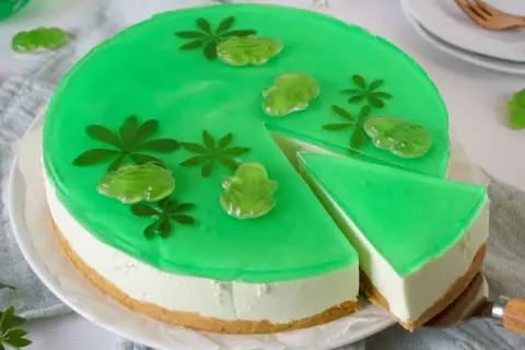 Grüne Philadelphia Torte Waldmeister mit Gummifröschen und Blättern verziert. Ein Stück wird gerade herausgeschnitten.