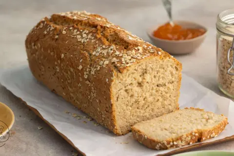 Frisch gebackenes Haferbrot, bestreut mit Haferflocken. Ein Glas Haferflocken und Marmelade daneben deuten auf ein leckeres Haferbrot Rezept hin.