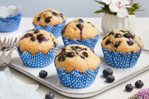 Fünf saftige, vegane Blaubeermuffins in blauen Papierförmchen auf einem Teller, garniert mit frischen Blaubeeren und einer Blume.