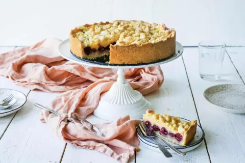 Ein saftiger Kirschkuchen mit Pudding und Streuseln auf einem Kuchenständer. Ein Stück ist bereits auf einem Teller angerichtet.