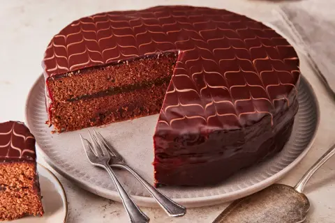 Eine glänzende Sacher Torte mit Schokoladenglasur und einem Stück Marmeladenfüllung. Gabeln liegen bereit.