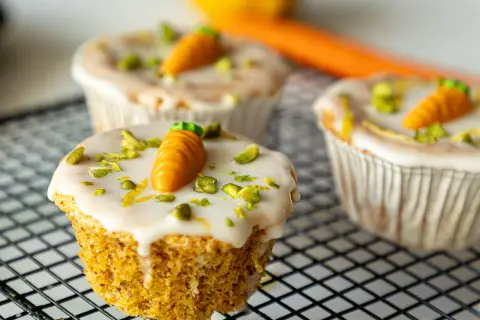 Drei leckere Karotten Muffins mit weißer Glasur, gehackten Pistazien und kleinen Marzipan-Karotten auf einem Gitter.