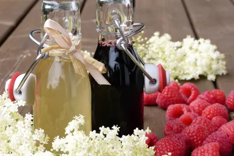 Zwei Flaschen mit selbstgemachtem Sirup, umgeben von Holunderblüten und Himbeeren. Perfekt zum Sirup einkochen.