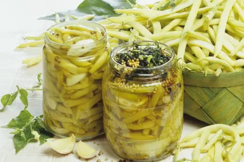 Zwei Gläser mit eingelegten Buschbohnen, daneben frische Bohnen und Knoblauch.