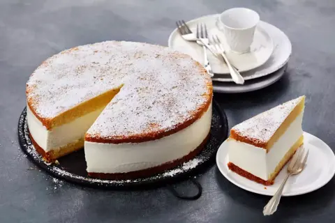 Eine köstliche Quark Sahne Torte, bestäubt mit Puderzucker, wartet auf Genießer. Ein Stück ist bereits auf einem Teller angerichtet.