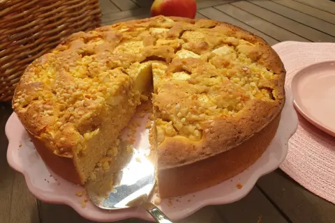 Ein saftiger apfelkuchen ohne zucker, bestreut mit Nüssen, wird auf einem rosa Teller präsentiert. Daneben liegt ein roter Apfel.