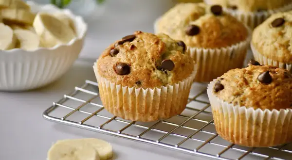 Frisch gebackene schoko bananen muffins mit Schokostückchen auf einem Kuchengitter. Daneben eine Schüssel mit Bananenscheiben.