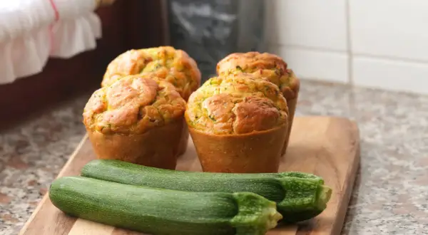 Vier goldbraune Zucchini-Muffins auf einem Holzbrett, daneben zwei frische Zucchini.