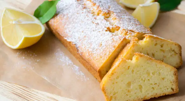 Saftiger veganer Zitronenkuchen, bestäubt mit Puderzucker, mit frischen Zitronenspalten und Blättern dekoriert.