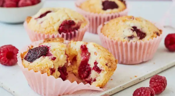 Saftige Himbeer Muffins in rosa Förmchen, einer aufgeschnitten, mit frischen Himbeeren garniert.