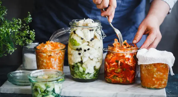 Kimchi selber machen im Glas: Verschiedene fermentierte Gemüse in Gläsern, darunter ein Glas mit rotem Kimchi, das gerade mit einer Gabel entnommen wird.