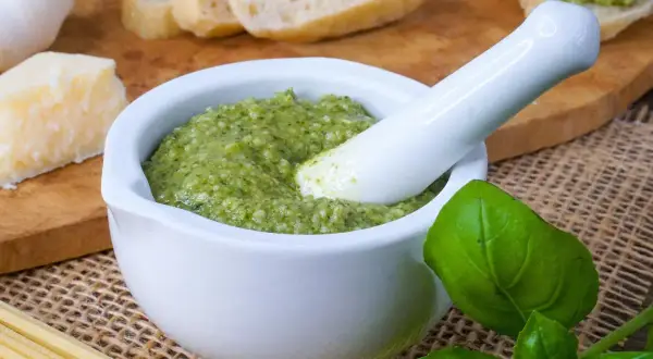 Frisches Pesto in einem Mörser, bereit zum Haltbarmachen mit Salz. Daneben liegen Brot, Parmesan, Knoblauch und Pasta.