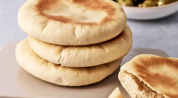 Gestapeltes pita brot mit einer Schüssel Oliven im Hintergrund. Ein Stück ist aufgeschnitten und zeigt die weiche Krume.