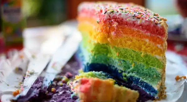 Ein Stück bunter regenbogen kuchen mit Streuseln, das auf einem Teller liegt. Daneben liegen zerbröselte Kuchenreste und ein Messer.