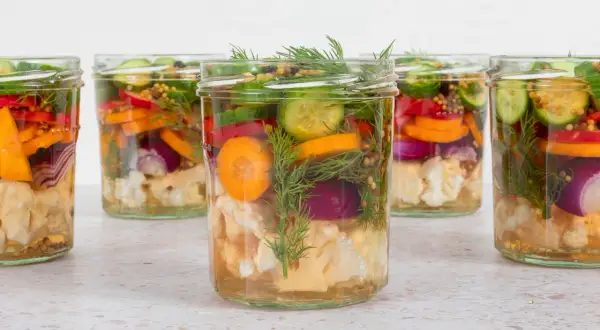 Sechs Gläser mit bunten Gemüsesorten wie Blumenkohl, Karotten, Gurken und Zwiebeln, bereit für mixed pickles selber machen.
